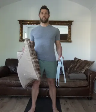 Kevin standing barefoot on a mat in front of a leather couch, holding a pillow in one hand and a strap in the other.