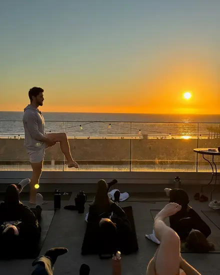 Kevin Cox balancing on one leg on a rooftop by the ocean at sunset, leading a workshop group lying on mats.